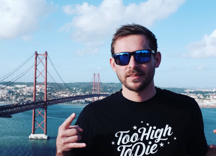 A man in a Too High To Die tee in front of Lisbon's 25 de Abril bridge