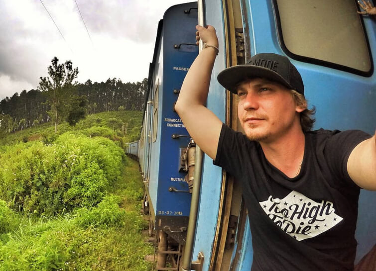 A traveler in a Too High To Die tee leaning out of a train in a tropical landscape