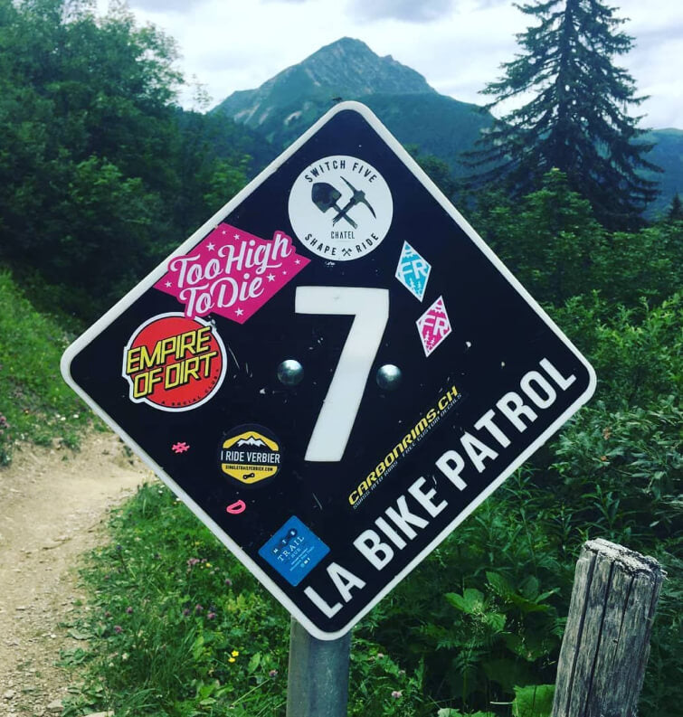 A Too High To Die sticker on a trail marker sign in an alpine bike park