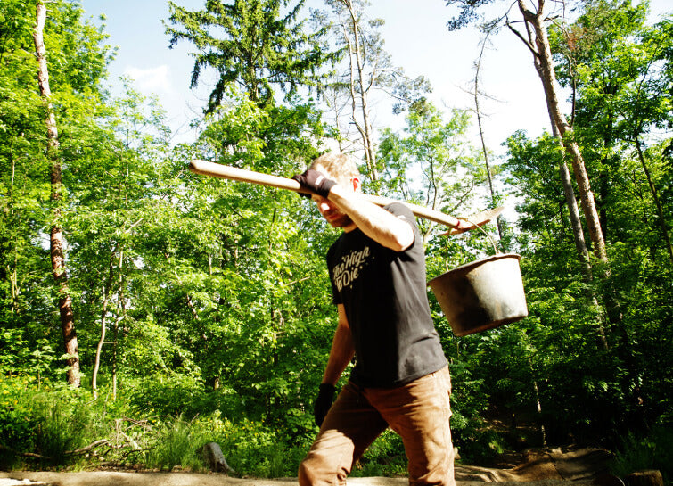 Trail building in the forest – a man in a Too High To Die tee carrying a mattock and bucket